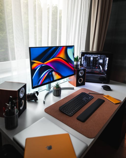 Escritorio de trabajo moderno y organizado con monitor de alta resolución, teclado mecánico, altavoces de madera y una torre de PC con iluminación blanca junto a una ventana.